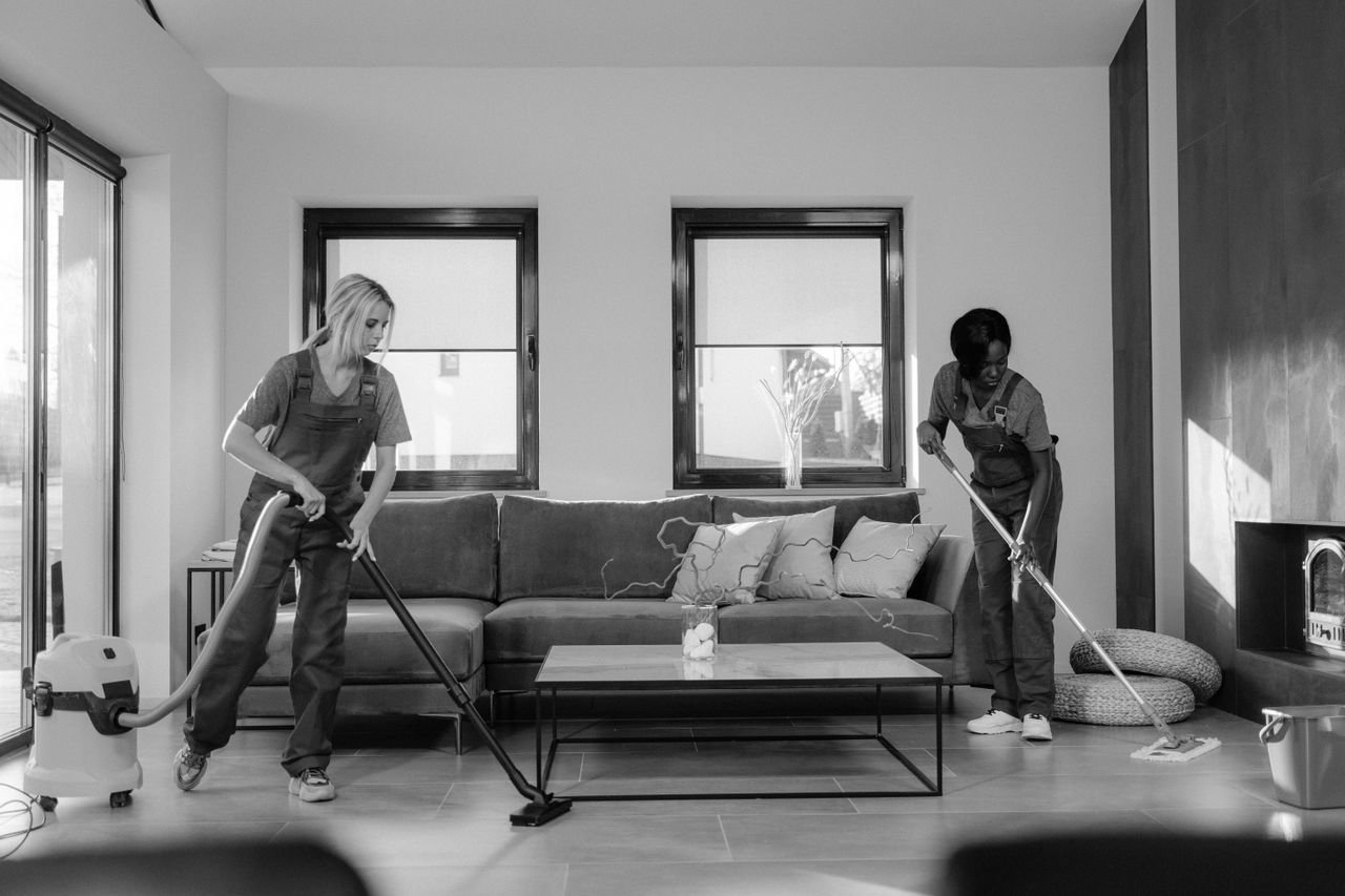 Mastering the First Impression: Your intriguing post title goes here Two women in work clothes cleaning a modern living room using a vacuum and mop.